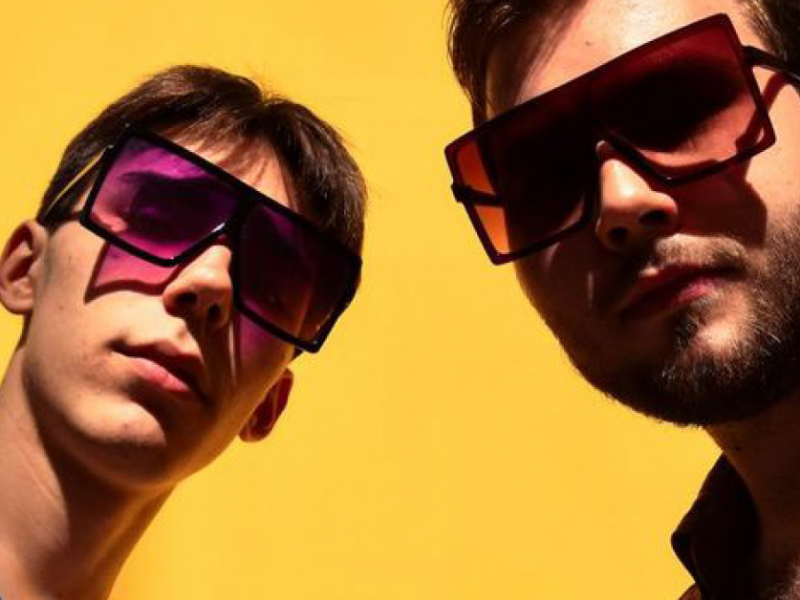 24.Avēnija - Music Duo: Two men, Ernests Vīgners and Kārlis Grīnbergs, standing with sunglasses against a vibrant yellow background, both looking straight to the camera.